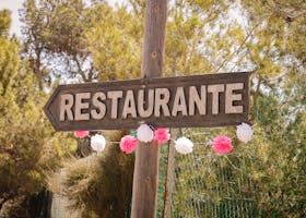 A rustic wooden restaurant sign in Spain, adorned with festive pink and white decorations amidst a natural outdoor setting.
